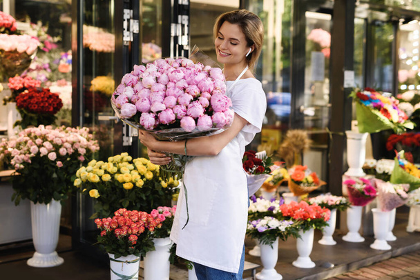 Young  woman florist with a heap of pink peony flowers in her little flower shop - Zdjęcie, obraz