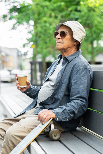 Stylish elderly man with a longboard sitting on the bench and drinking coffee in a city park - Фото, зображення