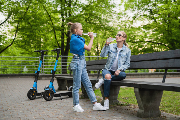 Happy mother and daughter with electric scooters are drinking lemonade in city park - Valokuva, kuva