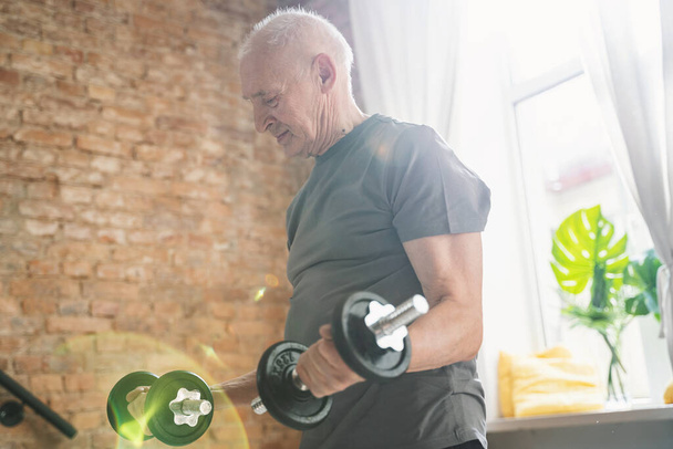 Ενεργός ηλικιωμένος άνδρας γυμνάζεται με αλτήρες κατά τη διάρκεια της προπόνησής του στο γυμναστήριο στο σπίτι - Φωτογραφία, εικόνα