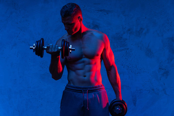 Young and muscular bodybuilder man exercising with dumbbells in colorful neon light - Foto, immagini