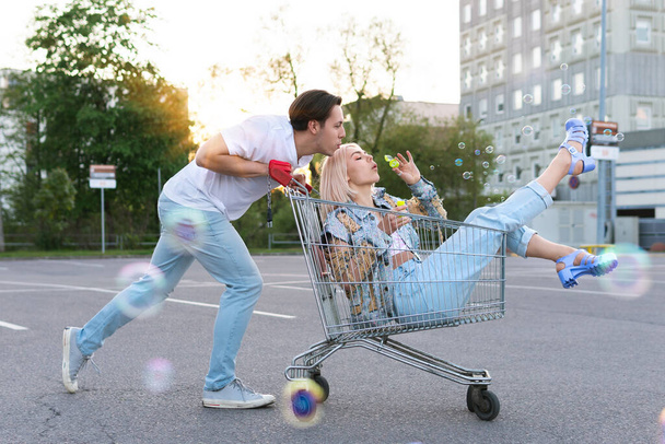 Happy young couple have fun with a shopping trolley and blowing bubbles on a supermarket parking - Foto, afbeelding