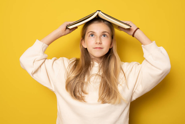 Beautiful girl with a book on her head isolated over yellow background. Education concept. - Photo, Image