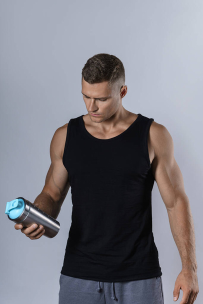 Muscular man wearing blank black tank top is drinking protein or other supplement from a metal shaker - Фото, зображення