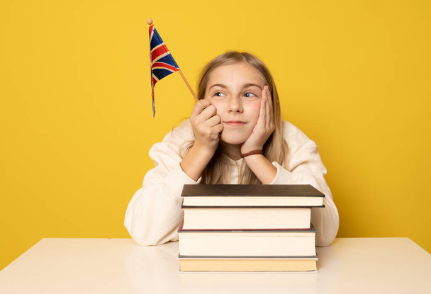 Beautiful smiling girl sitting on desk with pile of books and british flag over yellow background. Education concept. - Foto, Imagen