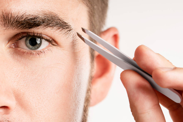 Close-up of male eye and tweezers for eyebrow grooming and shape correction - Foto, imagen