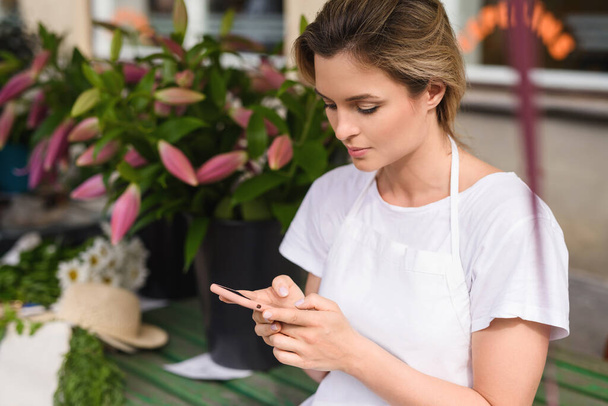 Young and beautiful woman florist using smartphone in her little flower shop - Фото, изображение