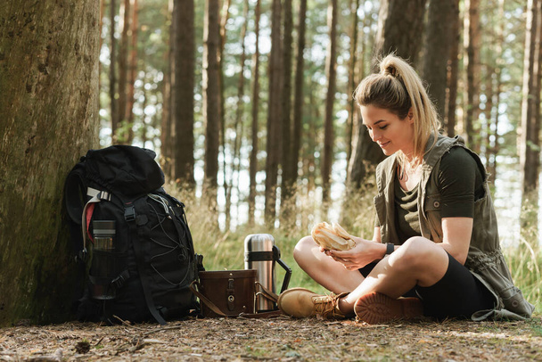 Young female hiker during small halt eating sandwich in green forest - Foto, afbeelding