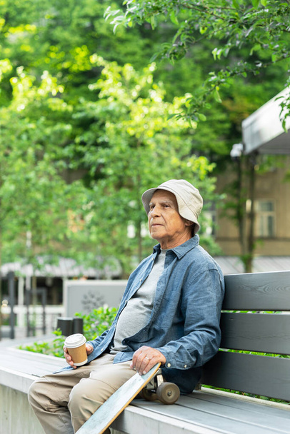 Stylish elderly man with a longboard sitting on the bench and drinking coffee in a city park - Foto, immagini