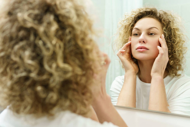 Young and beautiful woman with curly hair looking into the mirror in a bathroom - Photo, Image