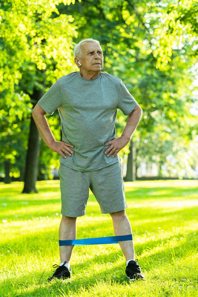 Active elderly man exercising with a rubber resistance band in green city park during his fitness workout - Foto, imagen