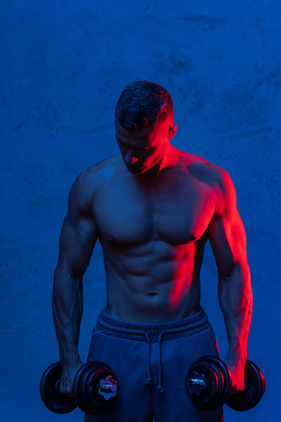 Young and muscular bodybuilder man exercising with dumbbells in colorful neon light - Фото, изображение