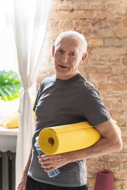 Active elderly man with a yellow mat and bottle of water is ready for his fitness workout at home  - Fotografie, Obrázek