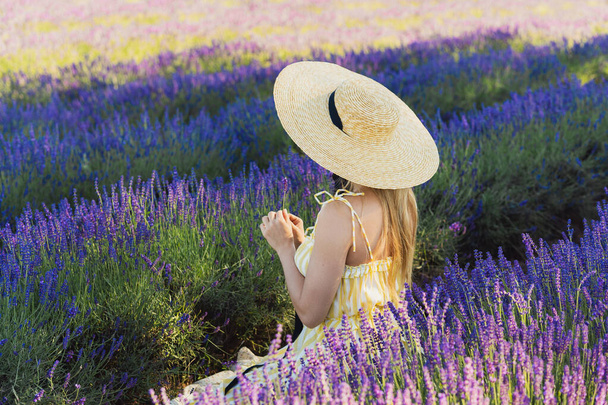 Beautiful young woman wearing hat sitting in the field full of lavender flowers - Fotoğraf, Görsel