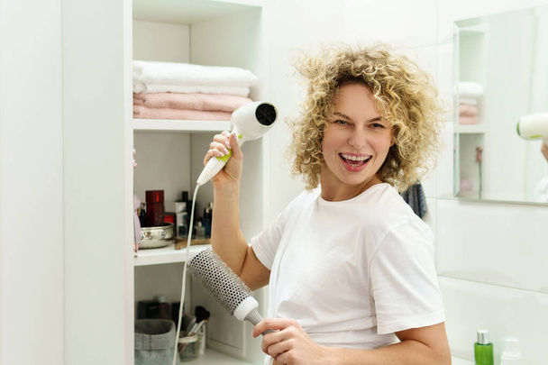 Young happy woman using hairdryer after for her curly hair in a bathroom - Foto, Imagem
