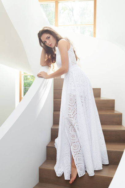 Portrait of gorgeous woman wearing beautiful white dress on a staircase inside her house - Fotó, kép