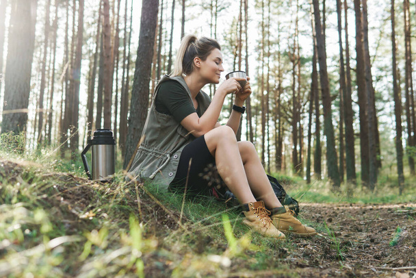 Young female hiker during small halt with a vacuum flask drinking hot coffee or tea in green forest - Φωτογραφία, εικόνα