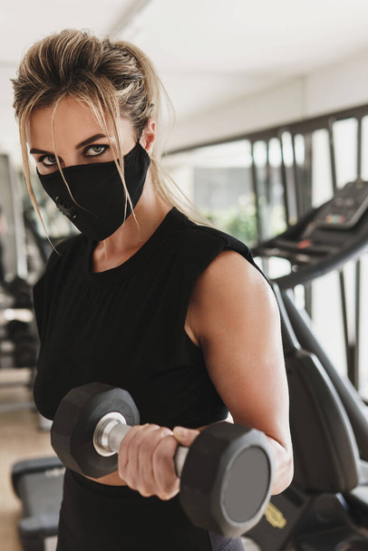Gym new normal. Young athletic woman wearing a prevention face mask during her fitness workout with a dumbbells. - Valokuva, kuva