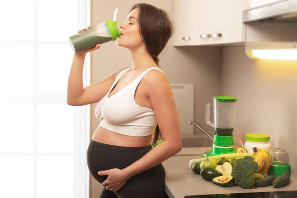 Young and happy pregnant woman drinking green protein cocktail at home - Foto, Imagem