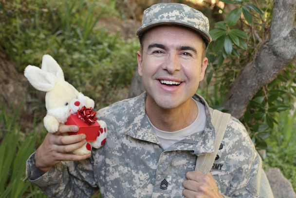 крупным планом портрет красивого молодого солдата с игрушечным кроликом и подарочной коробкой на открытом воздухе - Фото, изображение