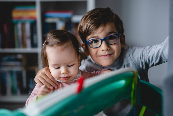 Happy small caucasian boy five years old wearing eyeglasses having fun playing with his baby sister playful siblings at home real people leisure concept childhood and bonding family - Photo, image