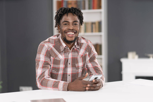 Uśmiechnięty przystojny Afroamerykanin używa smartfona sprawdzającego media społecznościowe lub aktualizującego informacje. Portret młodego mężczyzny trzymającego smartfona przy stole. Pojęcie mediów społecznościowych - Zdjęcie, obraz