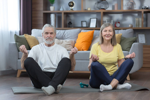 senior man and woman in the house doing yoga, family on a mat, in the lotus position looking at the camera together, morning exercise and fitness - Photo, Image