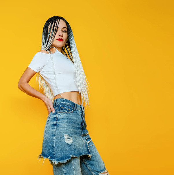 Bright photo of young woman on yellow background. Dreadlocks. White T-shirt and ripped jeans. - Photo, Image
