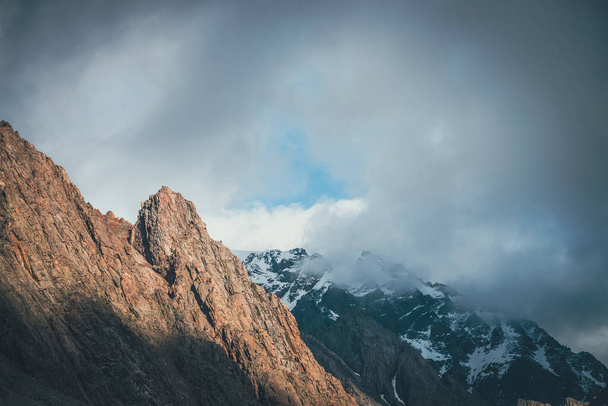 Niesamowity krajobraz z nasłonecznionym skalistym wierzchołkiem na tle wysokich śnieżnych gór w niskich chmurach. Atmosferyczna sceneria alpejska z ostrą skałą i gigantyczną śnieżną górą na zachmurzonym niebie. Malowniczy widok alpejski. - Zdjęcie, obraz
