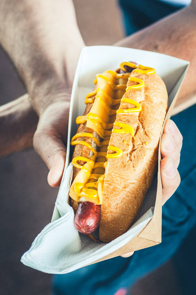 Freshly prepared cheesy hotdog in a paper box in a hand. Food delivery or lyfestyle concept. Close up. - 写真・画像
