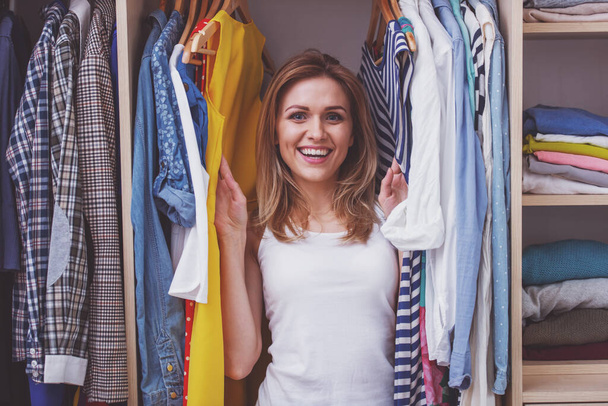 Beautiful girl is smiling and looking at camera while standing among clothes in her dressing room - Photo, Image