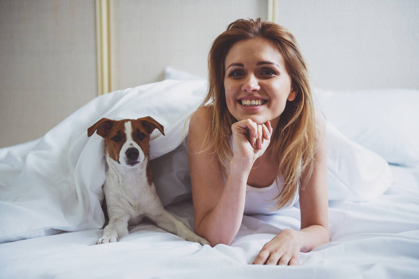 Güzel sarışın kız ve onun sevimli köpek kameraya yatakta bir battaniye ile örtülü yatan arıyoruz - Fotoğraf, Görsel