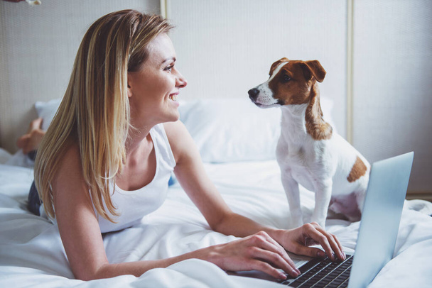 Beautiful girl in casual clothes is using a laptop, smiling and looking at her cute dog while lying on bed - 写真・画像