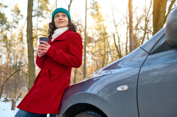 Rento kaunis kaukasialainen nainen matkustaa punaisessa takissa, lämmittää kätensä pitämällä lämpökuppia kuumalla teejuomalla tai kahvilla, istuu auton konepellillä ja nauttii talven viileästä säästä lumisessa mäntymetsässä. - Valokuva, kuva