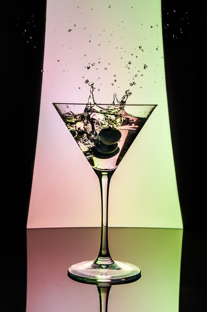 Ποτήρι κοκτέιλ με ελιές που πέφτουν μέσα και πιτσιλίζουν. Φόντο σε ροζ και πράσινο χρώμα πλαισιωμένο από θεατρικές κουρτίνες - Φωτογραφία, εικόνα