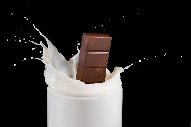 Barra de chocolate cayendo en una taza de leche y salpicaduras. Fotografía de alta velocidad sobre fondo negro - Foto, imagen