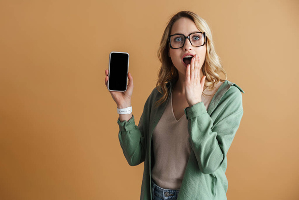 Shocked beautiful woman exclaiming while showing mobile phone isolated over beige wall - Fotoğraf, Görsel