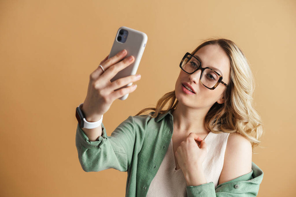 Pleased charming woman taking selfie photo on mobile phone isolated over beige wall - Фото, зображення
