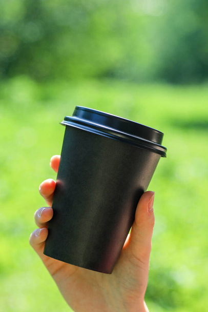 Black paper coffee cup in human hand in a sunny day on a green blurry background. Summer outdoor leisure. - Foto, Imagem