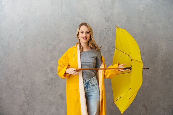Lovely cheerful young woman wearing waterproof coat holding umbrella isolated over gray background - Fotografie, Obrázek