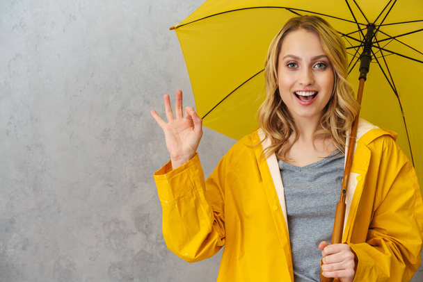Schöne fröhliche junge Frau in wasserdichtem Mantel hält Regenschirm isoliert über grauem Hintergrund, zeigt ok - Foto, Bild