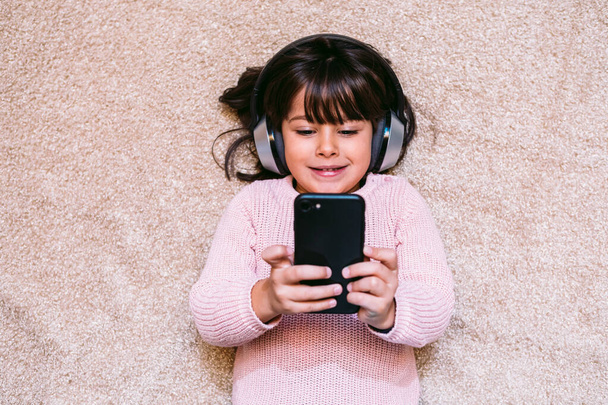 Kleines Mädchen im rosafarbenen Pullover, auf einem Teppich liegend, Musik hörend und Filme auf ihrem Handy schauend, mit kabellosen Kopfhörern. Konzept von Technik, kleinen Kindern und Handys. - Foto, Bild