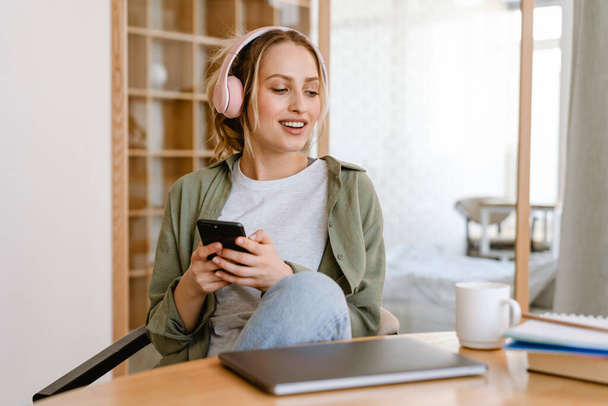 Nuori nainen hymyilee kuunnellessaan musiikkia matkapuhelin ja kuulokkeet kotona - Valokuva, kuva