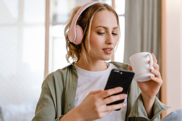 Young woman listening music with cellphone and headphones and drinking coffee at home - Fotó, kép