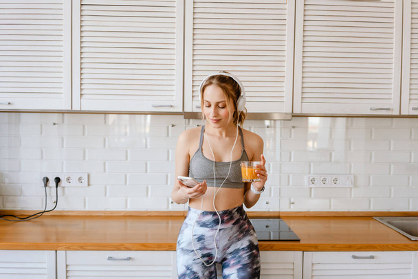 Smiling young white woman in sportswear drinking juice listening to music with headphone in the kitchen at home - Fotoğraf, Görsel