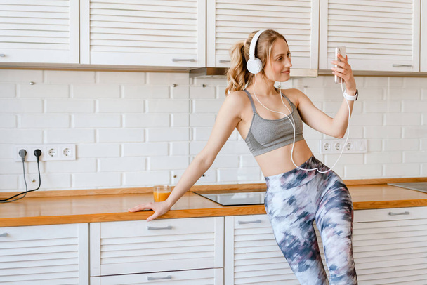Sorridente giovane donna bianca in abbigliamento sportivo bere succo di frutta ascoltare musica con le cuffie in cucina a casa prendendo un selfie - Foto, immagini