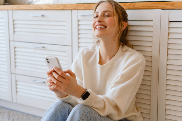 Young woman laughing and using mobile phone while sitting on floor at home - Foto, Imagen