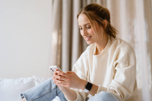 Young woman smiling while using cellphone in bedroom at home - Foto, Imagem