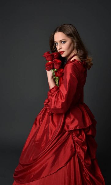  portrait of pretty female model with red hair wearing glamorous historical victorian red ballgown.  Posing with a moody dark background. - Photo, Image