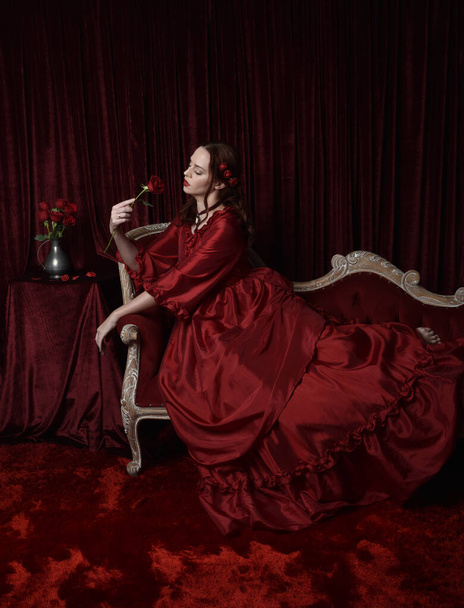  Porträt einer hübschen Frau mit roten Haaren in einem glamourösen historischen viktorianisch-roten Ballkleid. Posieren mit einem launischen dunklen Hintergrund. - Foto, Bild